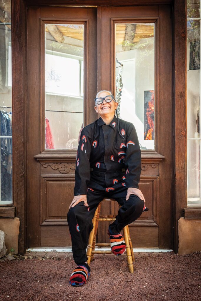Image of a woman sitting outside a shop on a stool smiling in an all black outfit with pops of color